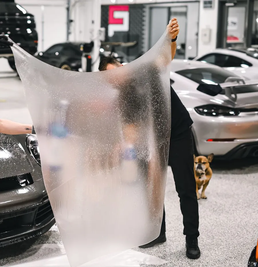 Two installers holding a large sheet of clear paint protection film in front of a sports car